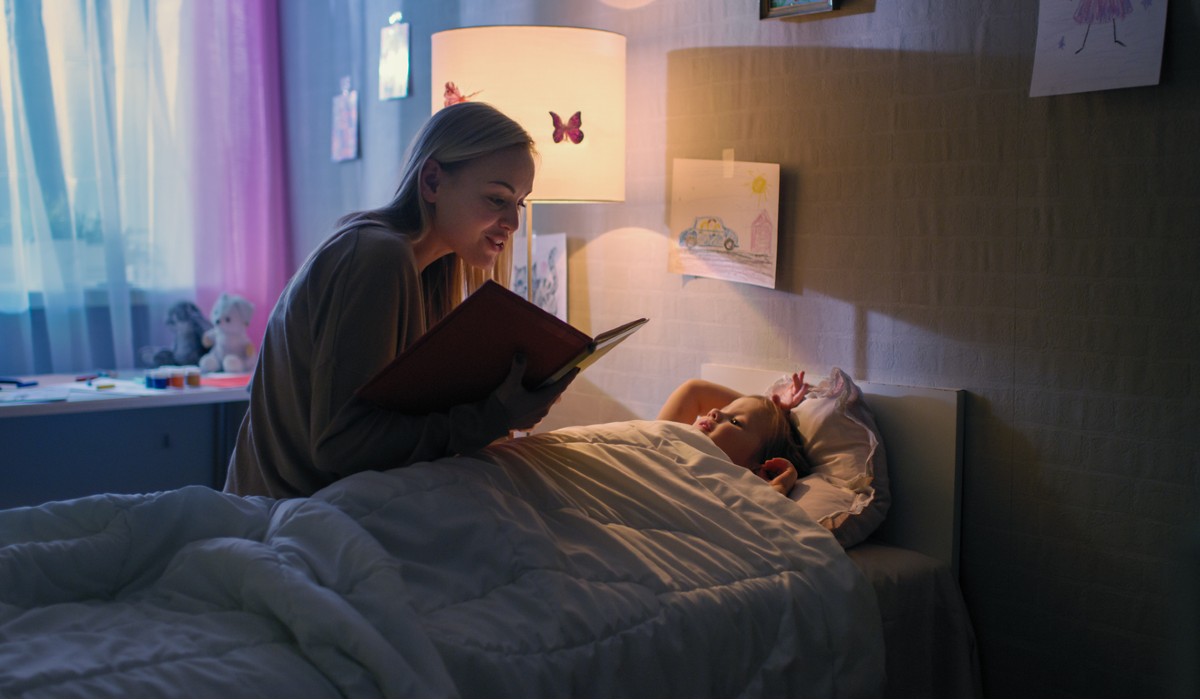 Young Loving Mother Reads Bedtime Stories to Her Little Beautiful Daughter who Goes to Sleep in Her Bed