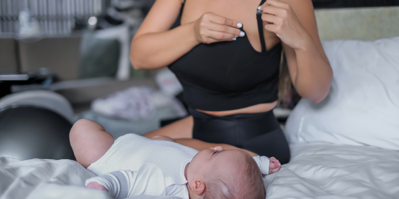 postpartum-clothes-for-new-moms mighty-moms Close-up shot of a mixed race mother unhooking the clasp on her breastfeeding black singlet bra while watching her newborn baby girl lying on the bed.