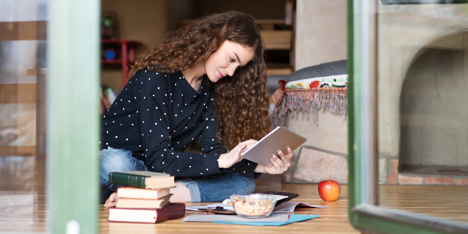 Beautiful teenage girl with long curly hair at home sitting on wooden floor, holding tablet, reading, studying, books and notebooks surrounding her.