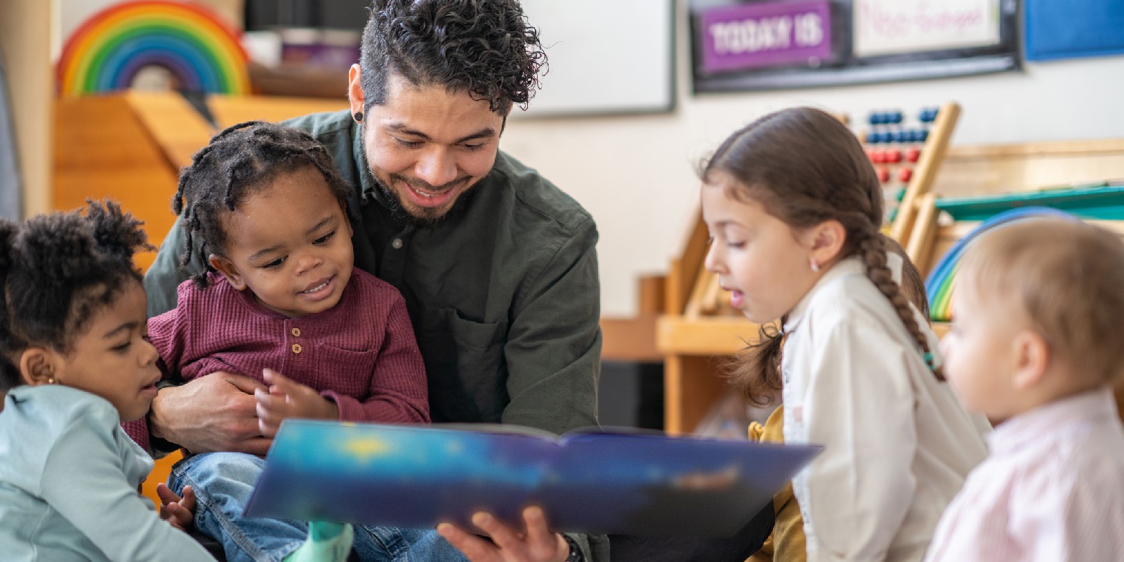 A male daycare educator of Hispanic decent, sits on the floor with a child on his lap and other toddlers scattered around him, as he reads them a story.