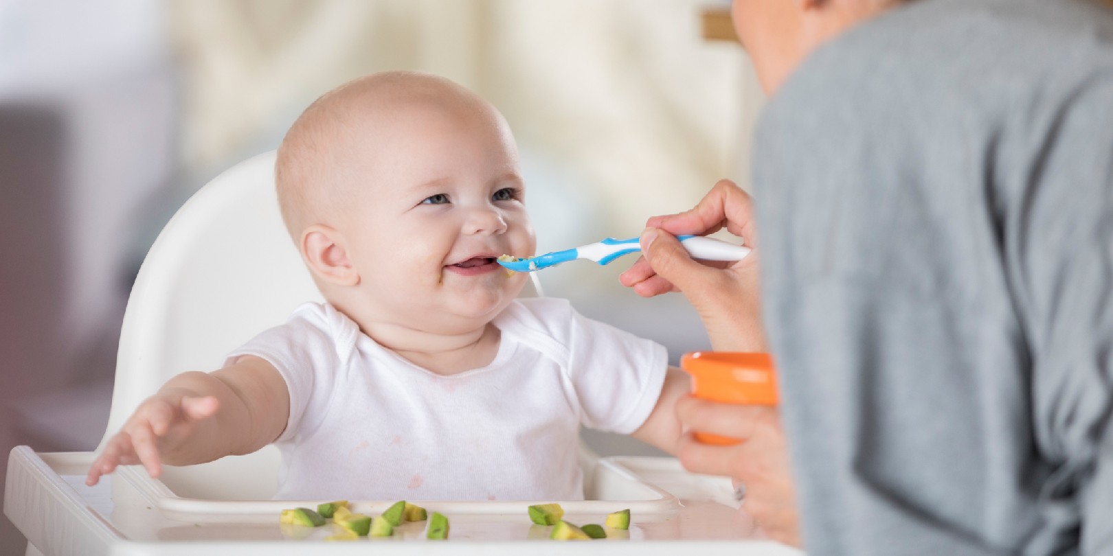 An adorable baby in a high chair laughs at her mother as she spoon feeds her. She pretends the spoon is a moving train using sound effects.