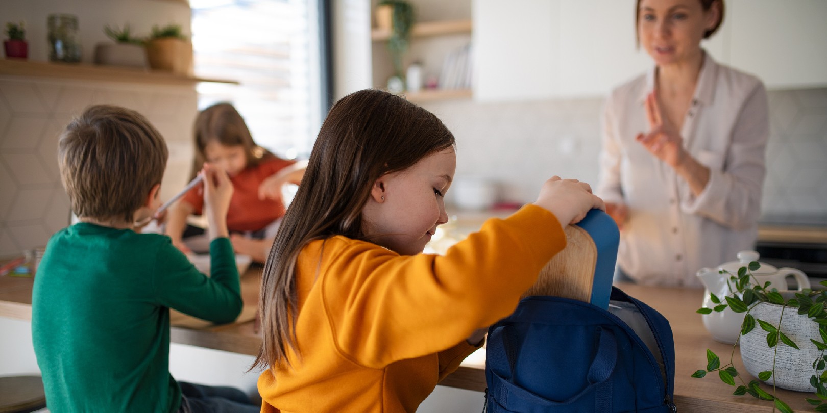 A little girl packing lunch box to backpack in kitchen at home.