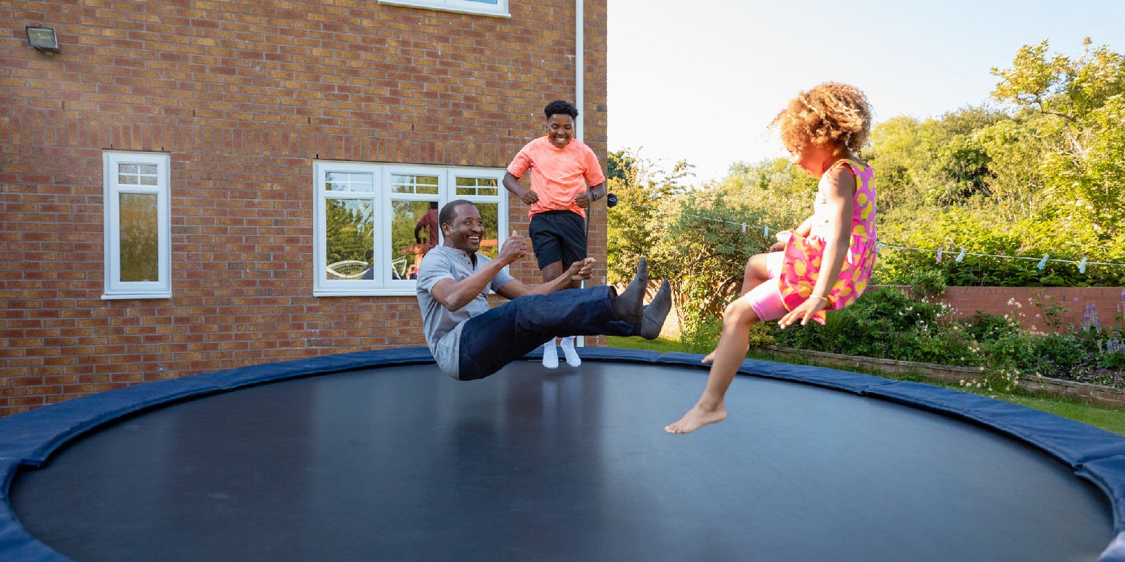 A young girl and her brother jumping on a trampoline outdoors in the back garden at home with their grandfather.