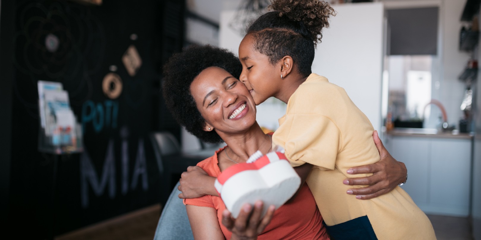 Young African American daughter surprising her mother at home with a nicely wrapped heart shaped giftbox on mother's day, making her feel important and bonding with her