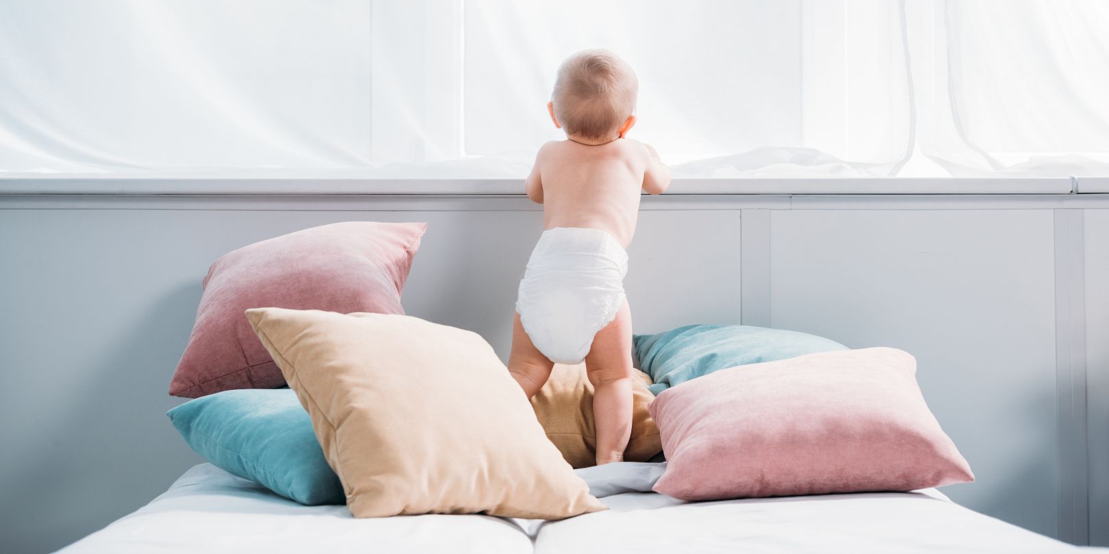 happy little baby in diaper standing on bed with lot of pillows and looking through window